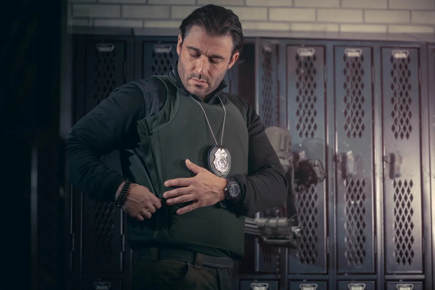 Un agente con placa de detective colgando en el cuello ajustando el cierre lateral de su chaleco antibalas discreto en una zona de casilleros operativos, mostrando la ergonomia de la marca.