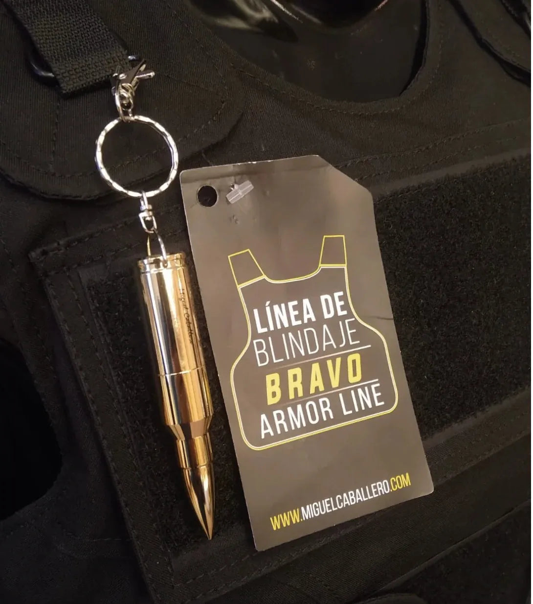 Primer plano de la etiqueta oficial de la Linea de Blindaje Bravo Armor Line de Miguel Caballero junto a un llavero en forma de bala dorada sobre un chaleco tactico negro.