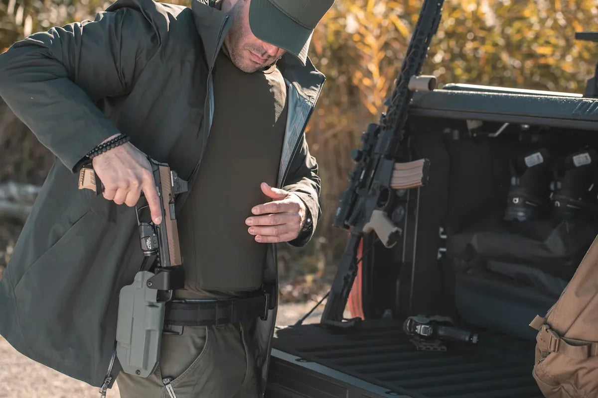 Un hombre con vestimenta tactica de exteriores desenfundando una pistola junto a la cajuela de un vehiculo con armamento, portando una prenda interior con proteccion balistica discreta de Miguel Caballero.