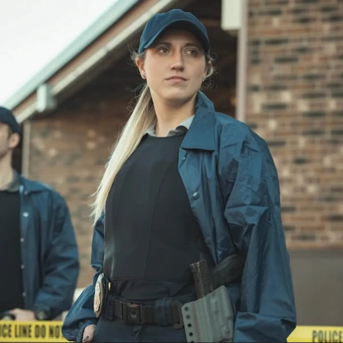 Una mujer oficial de seguridad portando una playera blindada negra debajo de una chamarra tactica azul, equipada con cinturon de servicio y placa oficial, demostrando proteccion balistica femenina de alta gama.