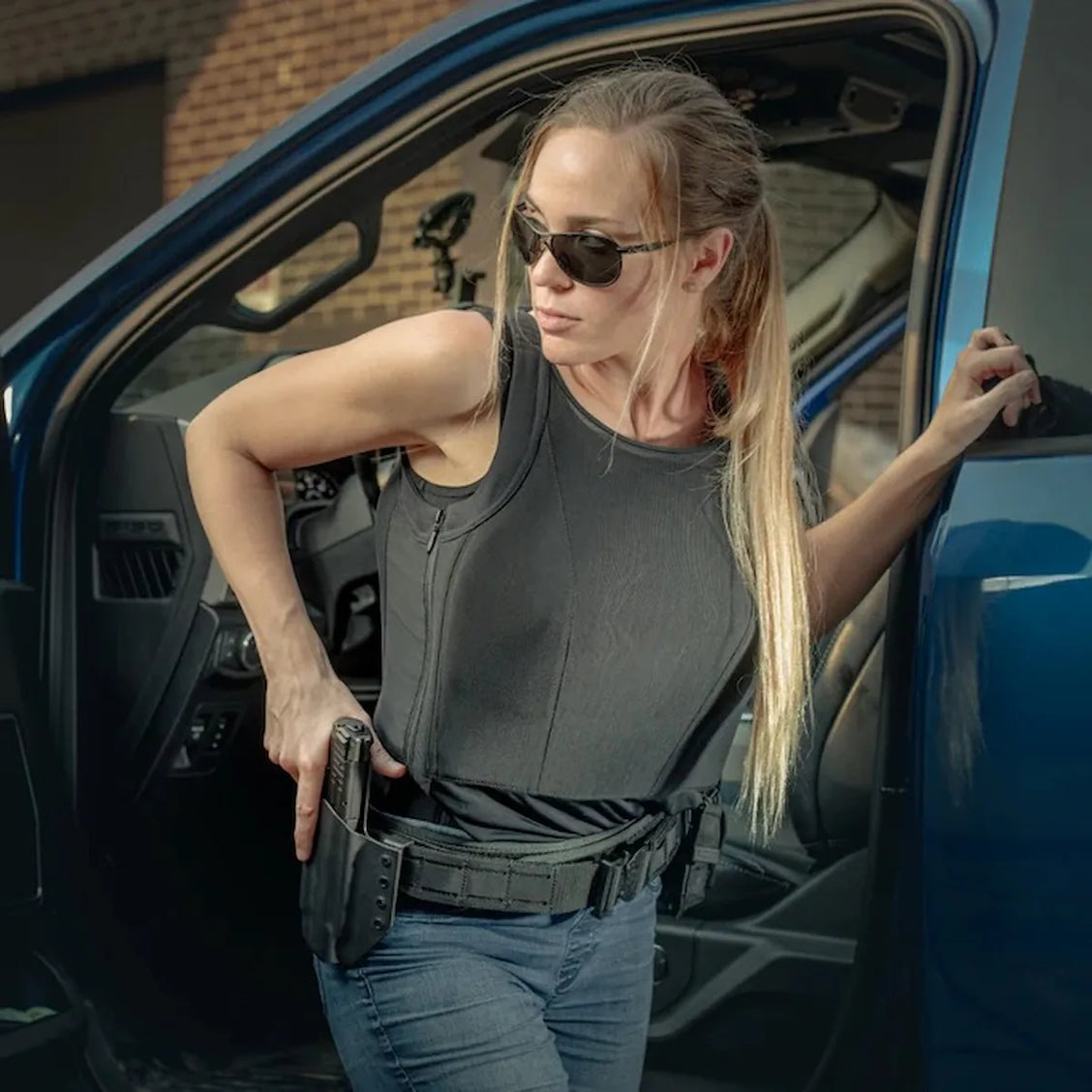 Primer plano de una mujer en accion saliendo de una camioneta azul, vistiendo la playera blindada femenina tipo tank top color negro de Miguel Caballero con equipo de extraccion rapida.